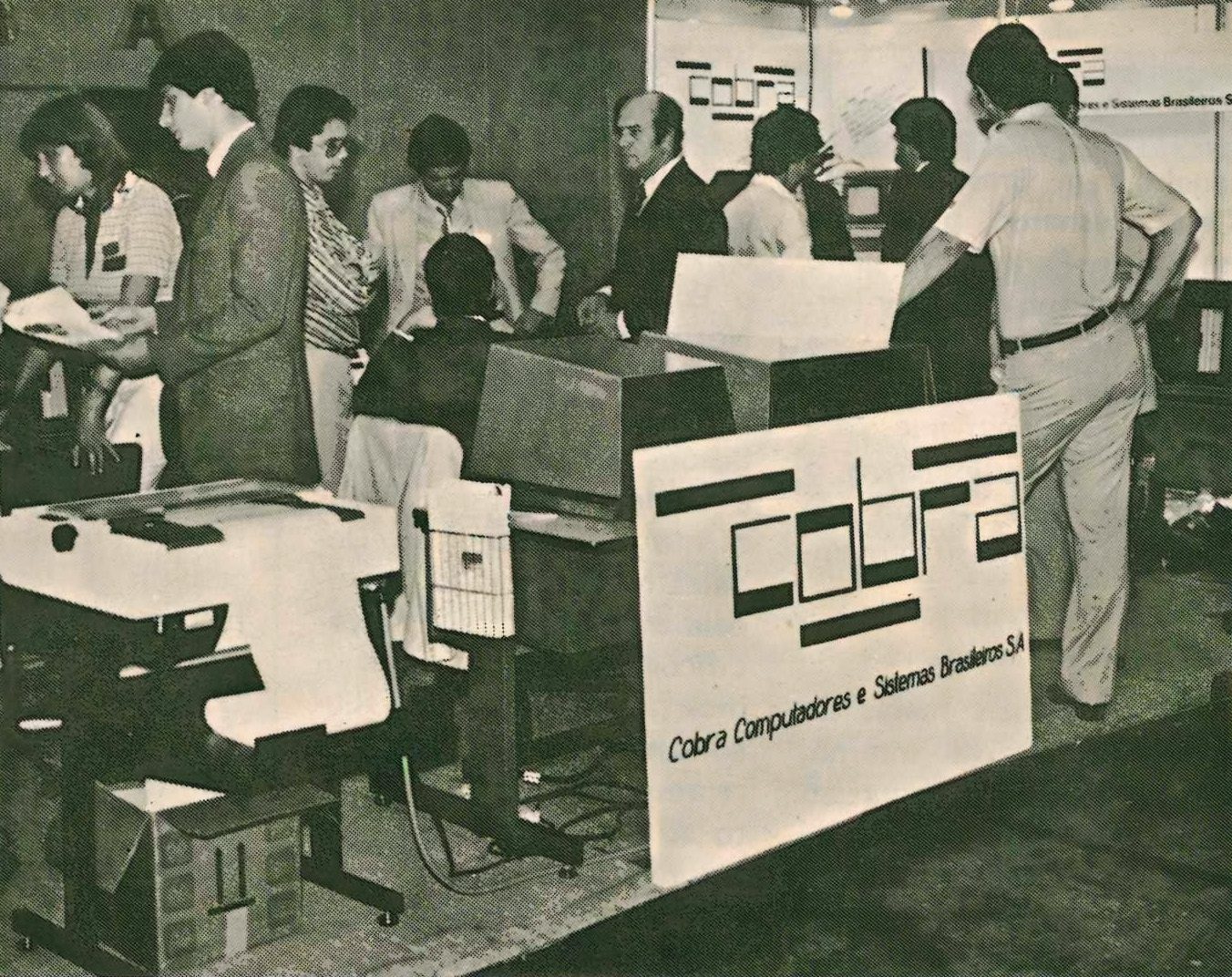 Foto de época, em tom envelhecido, de um estande da COBRA Computadores, com pessoas reunidas ao redor de um terminal e o logo da empresa em destaque.