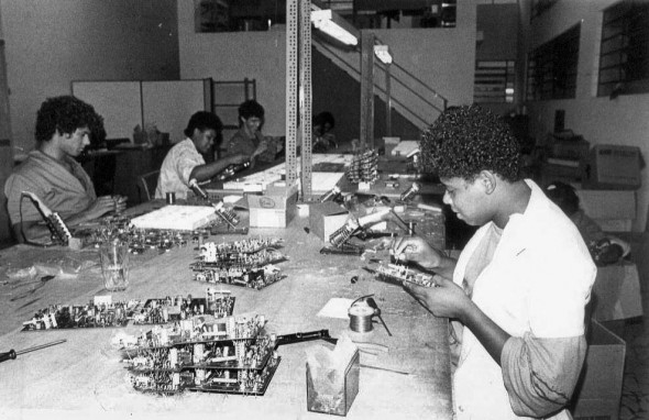 Foto em preto e branco de operárias em uma linha de montagem de fábrica, soldando e montando placas de circuito.