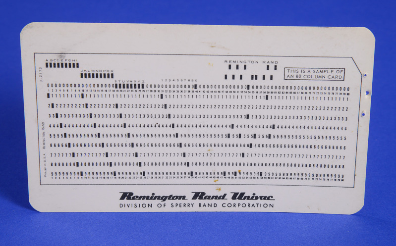Foto de um cartão perfurado branco da Remington Rand UNIVAC, mostrando fileiras de números, letras e padrões de furos que representam dados.