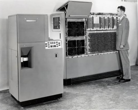 Foto em preto e branco de um homem de terno ao lado do computador UNIVAC 120, uma máquina de grande porte que ocupa uma sala.